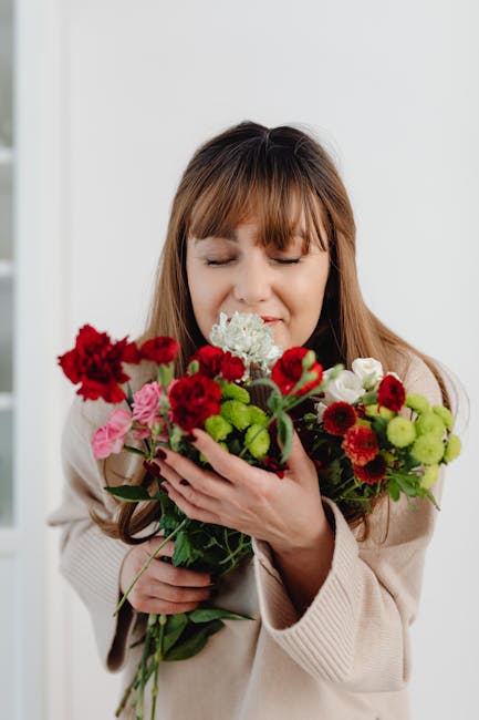 A woman with light brown hair and bangs, wearing a beige coat, is holding a large, vibrant bouquet of fresh flowers. The bouquet includes red carnations, pink roses, white lisianthus, green chrysanthe