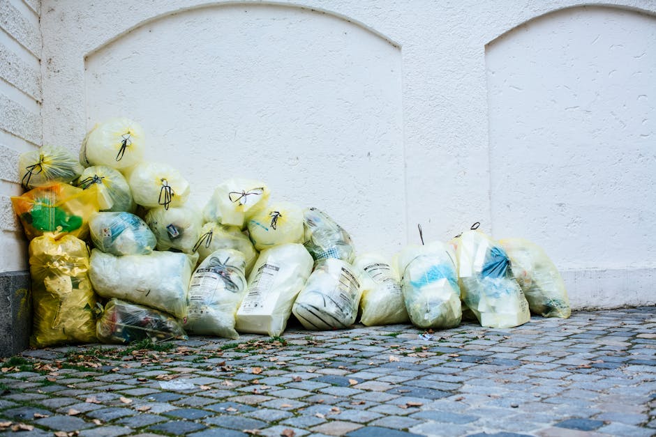 A pile of yellow, white, and blue plastic garbage bags filled with household waste, including some visible packaging and paper, placed on a cobbled street next to a white wall and a brick corner. The 