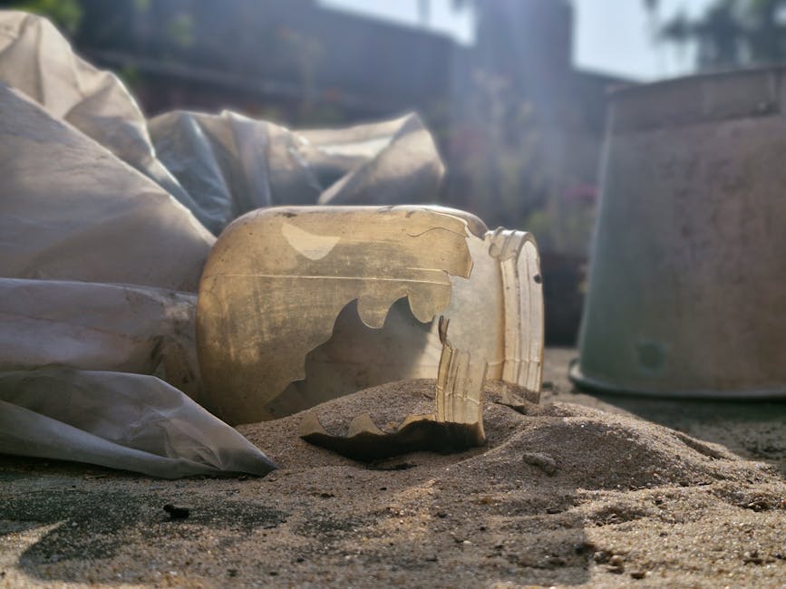 A broken ceramic skull-shaped object with visible cracks and missing pieces, resting amidst sand and soil on an outdoor surface. Surrounding the sculpture are discarded plastic wrapping and a silver c