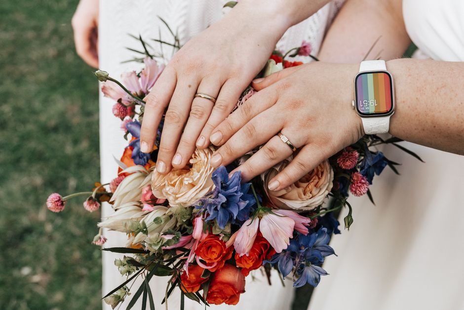 A close-up of two hands holding a vibrant floral bouquet, featuring large peach roses, blue delphiniums, bright orange and pink ranunculus, and small pink and purple blooms, wrapped in natural tissue 