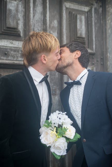 A romantic moment captured of two men dressed in formal tuxedos and bow ties sharing a kiss outside a building with dark, textured stone walls. One man has light blond hair, while the other has dark h