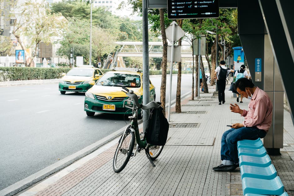 A man wearing a light pink shirt and dark jeans, sitting on a blue plastic bench at a bus stop on Turnpike Lane in Harringay, looking down at his phone. Next to him, a black bicycle with a front baske