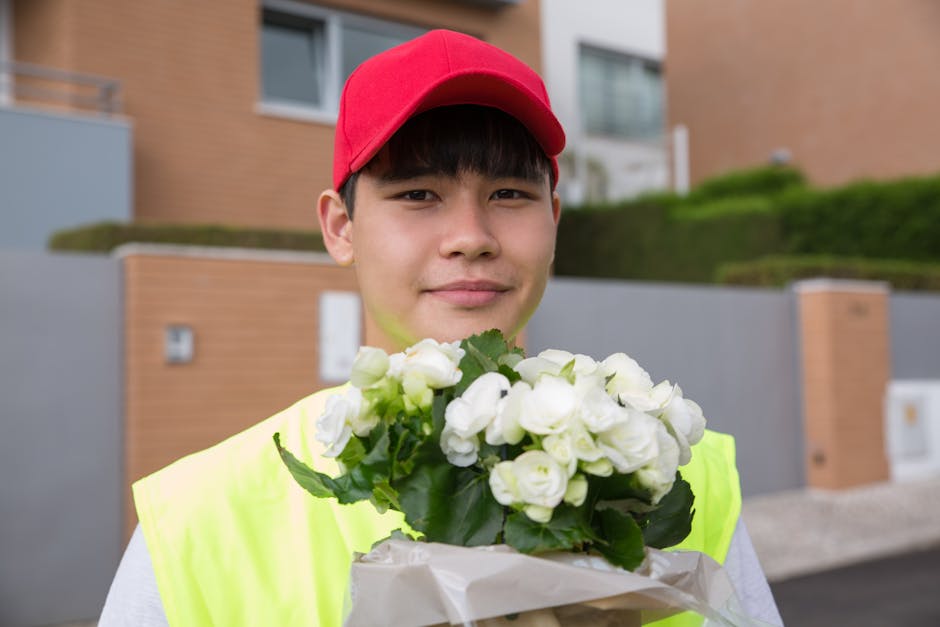 A young male flower delivery courier wearing a bright yellow high-visibility vest and a red cap, holding a bouquet of white freesias wrapped in white paper, with green leaves visible. The courier is s