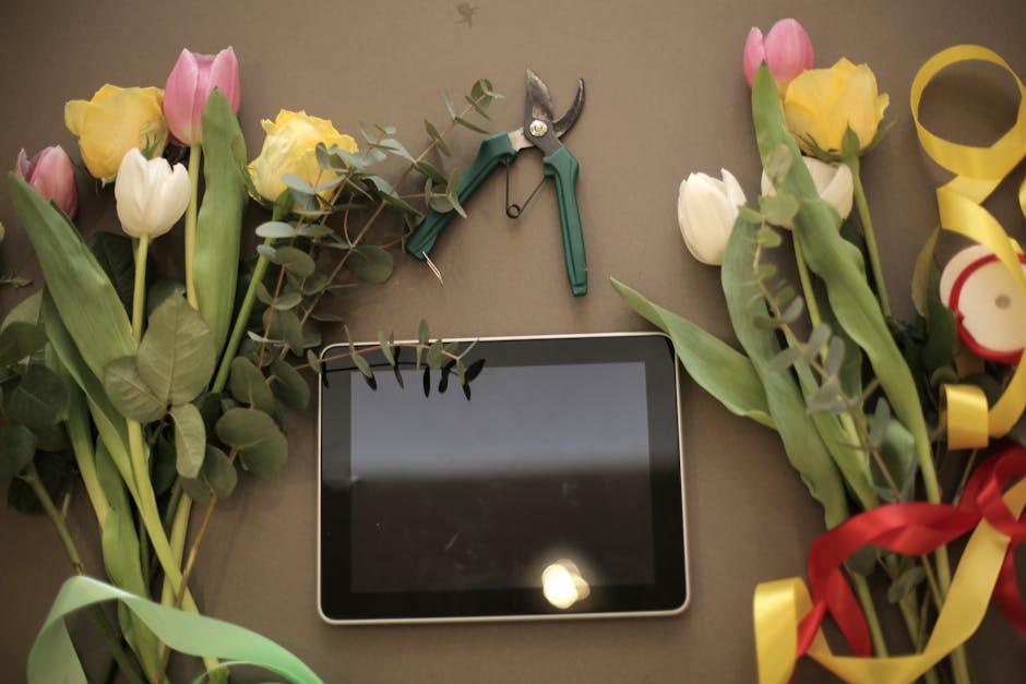 A flat-lay arrangement of fresh tulips and roses in soft pastel yellow, pink, and white, with green foliage, placed around a digital tablet on a neutral surface. The flowers are in various stages of b