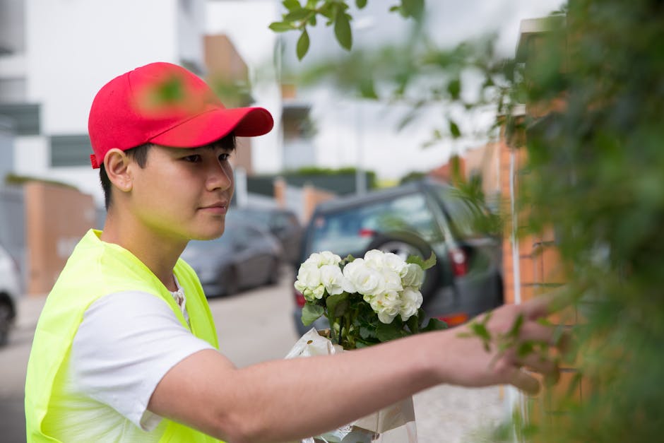A young delivery person wearing a red cap and a neon yellow safety vest is placing a bouquet of fresh white hydrangea flowers wrapped in white paper against a brick wall with green foliage in the fore